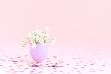 Obraz na płótnie Canvas Purple easter eggshell with white gypsophila flowers on pink background with multicolor confetti. Egg is symbol of celebration of a religious holiday among Catholics, Christians, Protestants