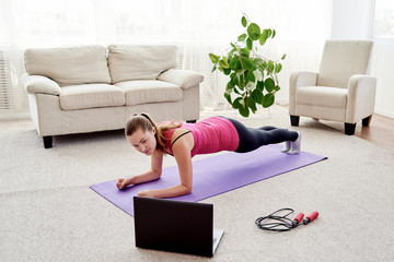 Beautiful young woman in sportwear doing plank exercise in living room at home, watching videos on laptop computer and repeating online instructions by coach, copy space. Full length body