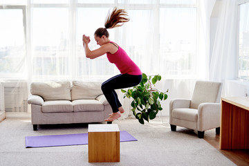 Young woman doing box jump exercise in living room at home, copy space. Jumping squats. Sport, healthy lifestyle concept