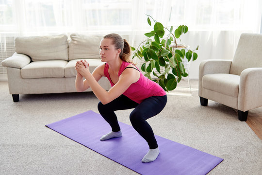 Young attractive woman in pink sportswear doing squats exercise at home, stretching legs. Full length portrait. Healthy lifestyle concept