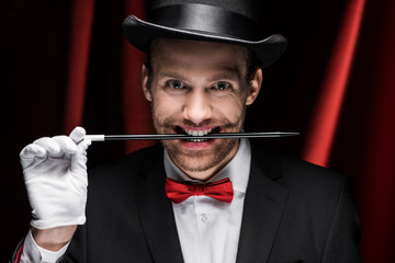 professional scary magician in suit and hat holding wand in teeth in circus with red curtains