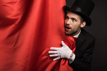 handsome emotional magician in suit and hat in circus with red curtains - obrazy, fototapety, plakaty