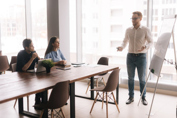 Businessman doing presentation, using whiteboard, explaining to colleagues in modern office meeting against large panoramic window. Concept of office life.