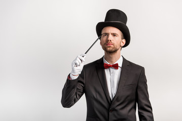 pensive magician in suit and hat holding wand, isolated on grey