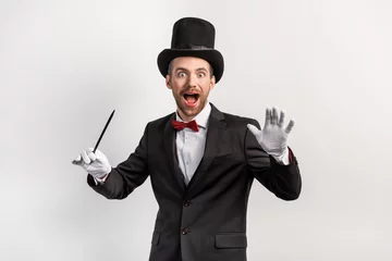 Fotobehang excited magician in suit and hat holding wand, isolated on grey © LIGHTFIELD STUDIOS