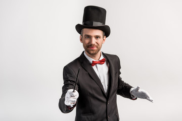 positive magician in suit and hat holding wand, isolated on grey