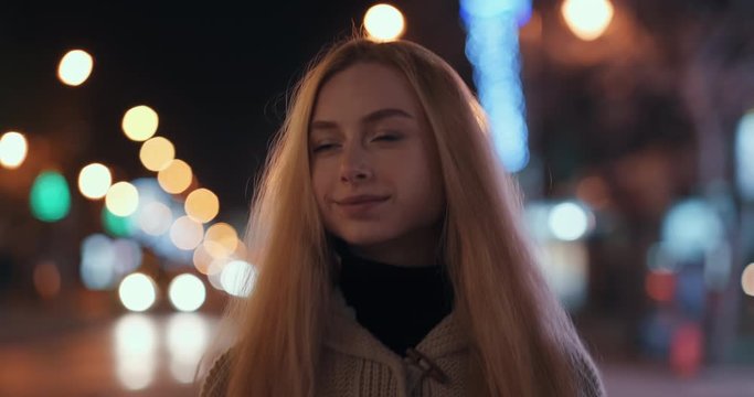 Portrait of happy blonde girl with long hair stands and smiles, looks at camera against of passing cars and bright bokeh of city lights in evening in winter after walk. Emotions of people. Lifestyle