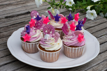 Cupcakes for birthday party with stars decorations on a ceramic plate in a rustic style. Vintage wooden background with flowers. Cakes decorated with white and pink cream, candies and chocolate.