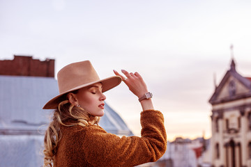 Outdoor close up portrait of young attractive elegant woman wearing beige wide brim hat, stylish wrist watch, brown faux fur coat, posing in European city. Copy, empty space for text