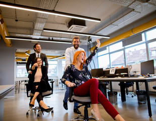 Young business people in smart casual wear having fun while racing on office chairs and smiling