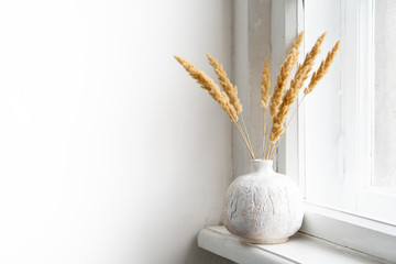 White textured hand-made ceramic vase with dry ears on windowsill - obrazy, fototapety, plakaty