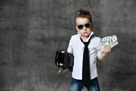 Serious Boy Little Businessman In White Shirt, Jeans, Tie And Sunglasses Standing And Holding Toy Car Present And Dollars Cash