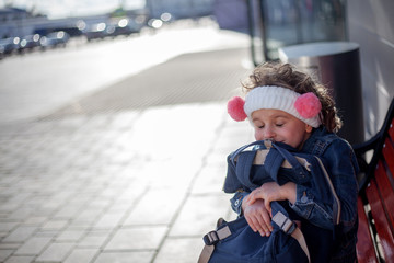 Happy kid sit on bench and hugging the backpack at the street of the city against the sunlight. Fashion chid wearing denim urban wears