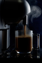  Coffee machine makes latte in a glass cup on a dark background. Cappuccino, cooking, cup, glass, morning, breakfast, steam, hot drink, brewing,