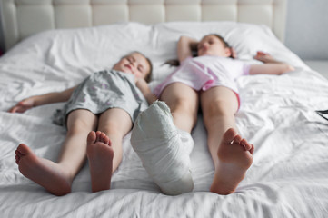Two girls are lying on the bed. The older sister has a leg in a cast. Medical concept