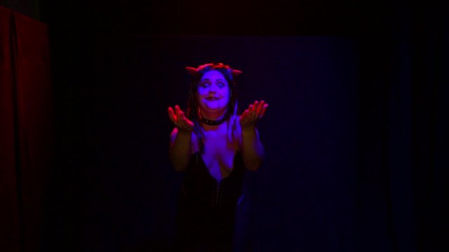 A woman in the form of a devil with an ominous smile dances in red lighting on a black background.