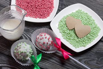 Cake pops in white glazed chocolate with green and pink sprinkles. A green and pink bow is tied on sticks. Nearby melted chocolate and topping in plates. Against the background of brushed pine boards