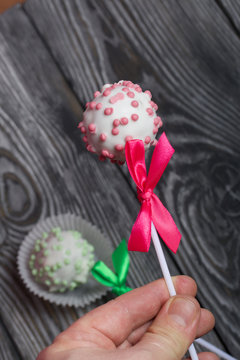 A man holds cake pops in white glazed chocolate with pink sprinkles. A green and pink bow is tied on a stick. Against the background of brushed pine boards painted in black and white.