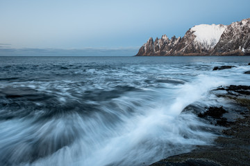 Waves in winter landscape crushing on dramtic rocks with snow covered Oksen mountains in...