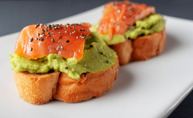Salmon bruschettas with avocado on a black background, decorated with chia seeds.