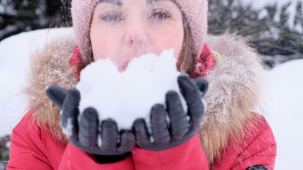 Smiling young woman in red jacket throwing snow in the air on a sunny winter day. Woman blows snow from her hands