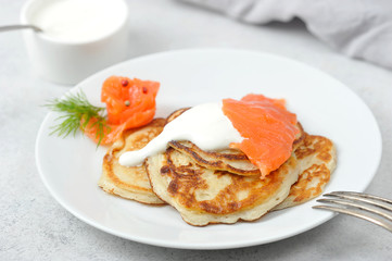 Fritters with salted salmon and sour cream.  Fork on the edge of the plate.  Light background.  Close-up.