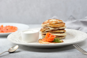 Pancakes with salted salmon.  Fritters served with salmon and sour cream.  Cutlery and a napkin complete the table.  Light background.  Close-up.