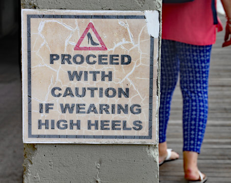 Sign to warn of the danger of wearing high heels. A lady with flat heels walks on the boardwalk