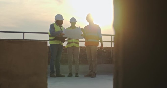Caucasian woman boss with laptop computer standing on the sunset on the roof with two male multiethnic builders.