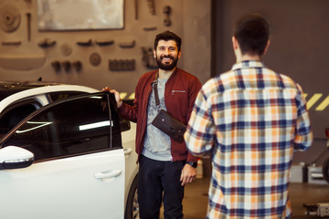 smiling male customer standing at his car and looking at the camera with mechanic in foreground