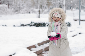 Beautiful little girl with a cat face in bright and bright clothes holding a snowball against the background of a snow covered park