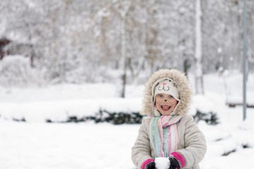 Fototapeta na wymiar Beautiful little girl with a cat face in bright and bright clothes holding a snowball against the background of a snow-covered park