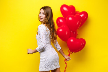 Obraz na płótnie Canvas beautiful young woman in white dress holding heart shaped balloons posing on yellow background