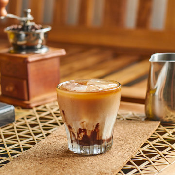 High angle of transparent glass of yummy coffee with fresh milk and ice standing on cork coaster beside small metal coffee pot and vintage grinder on brown wooden table at home
