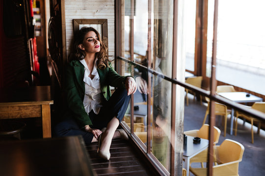 From above of pensive trendy woman sitting with crossed legs and looking through window of modern cafe