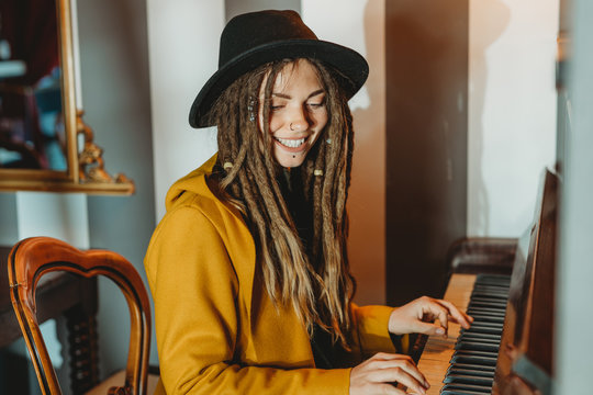 Side view of happy hipster female with dreadlocks wearing yellow coat and black hat playing piano while sitting in retro styled room