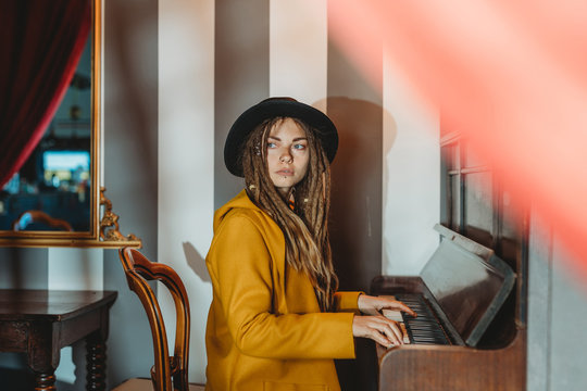 Side view of serious hipster female with dreadlocks wearing yellow coat and black hat playing piano while sitting in retro styled room