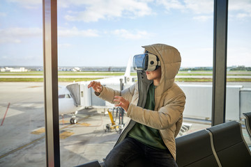 Preteen boy in hooded outwear and VR goggles gesturing and exploring virtual reality while sitting near window in modern airport