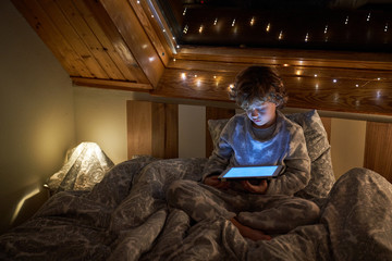 Little boy in pajama browsing tablet while lying in bed near window decorated with fairy lights at night in cozy room
