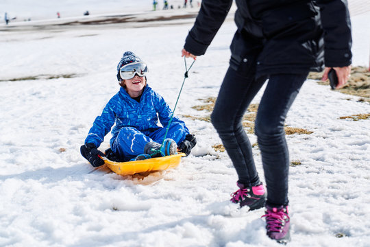 Little boy in warm sport clothes and snowboard goggles sitting on sled and laughing while cropped anonymous woman pulling sled and looking at boy in Candanchu, Huesca, Spain