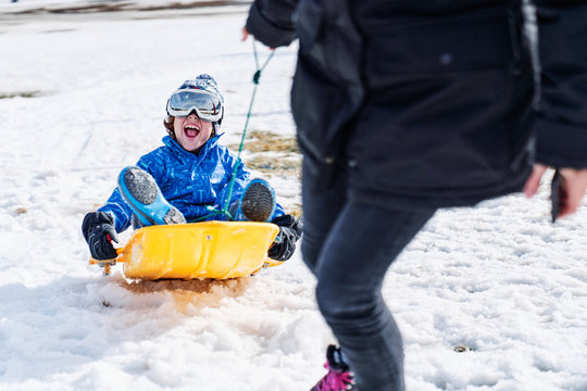 Little boy in warm sport clothes and snowboard goggles sitting on sled and laughing while cropped anonymous woman pulling sled and looking at boy in Candanchu, Huesca, Spain