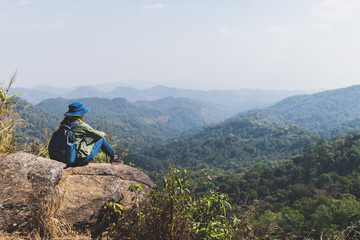 The back view of an asian woman traveler backpack and wearing a jean hat Sitting on the edge of a cliff in the forest, Thailand