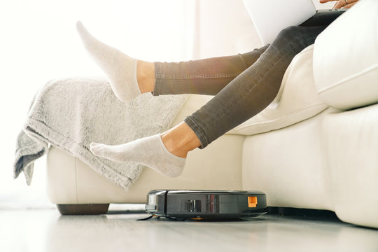 Side view of crop female in jeans and socks sitting on white sofa in room with laminate floor and putting feet on robotic vacuum cleaner