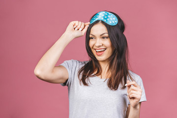 Waist up portrait of cheerful smiling girl in sleeping mask. Attractive funny female in stylish pajama standing and looking away. Isolated on pink background.