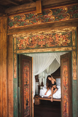 Obraz na płótnie Canvas Slim tan woman waking up and enjoying morning coffee in traditional wooden asian bed with white canopy. Wearing black lingerie. View through door of traditional balinese house. Bali vacations. 