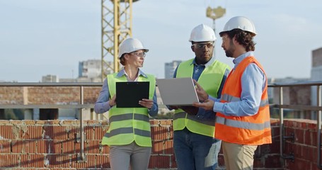 Three young mixed-races builders and architects, two men and woman standing together at the constructing site with laptop computer and tablet device and discussing work process.