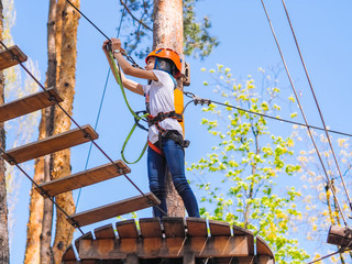 Teenager  in orange helmet climbing in trees on forest adventure park. Girl walk on rope cables and  high suspension bridge in adventure  summer city park.  Extreme sport equipment helmet and carabine