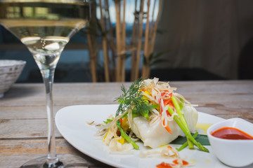 White fish seabass fillet with vegetables and white wine on a wooden table in a restaurant with decor. Tasty and healthy food. A cozy evening in a restaurant during a date. Beautiful table setting.