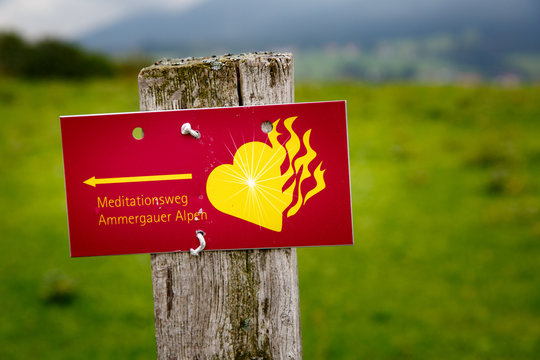 red sign in German: Ammergau Alps meditation trail. With a yellow flaming heart