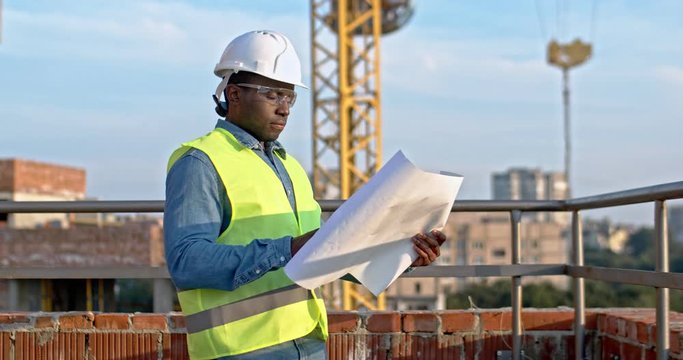 Portrait shot of the African American man constructor in the hardhat and goggles holding plans and drafts and studying them while standing outdoors at the building site.